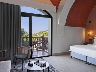 Helles Hotelzimmer mit Doppelbett, Stühlen, Tisch, Balkon und Blick auf grüne Hügel.
