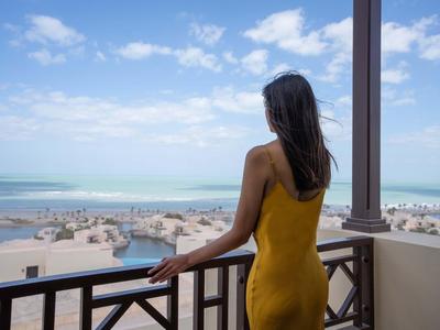 Frau im gelben Kleid blickt vom Balkon auf Strand, Meer und blauen Himmel mit Wolken.