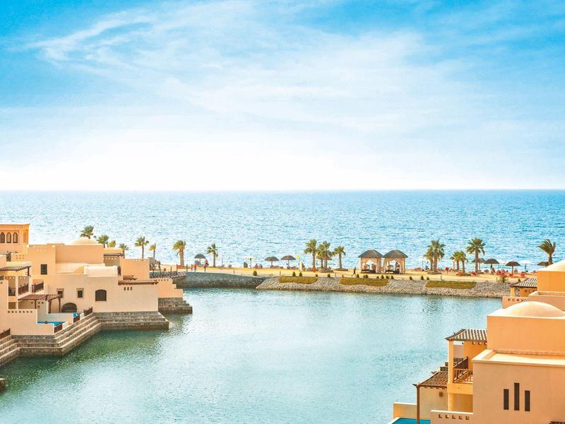 Blick auf türkisfarbenes Wasser mit beigen Gebäuden am Meer und blauem Himmel im Hintergrund.