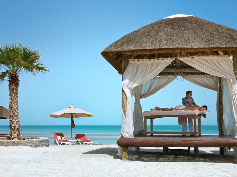 Strand mit Palmen, Liegestühlen, Sonnenschirmen und Massagetisch unter Strohdach.