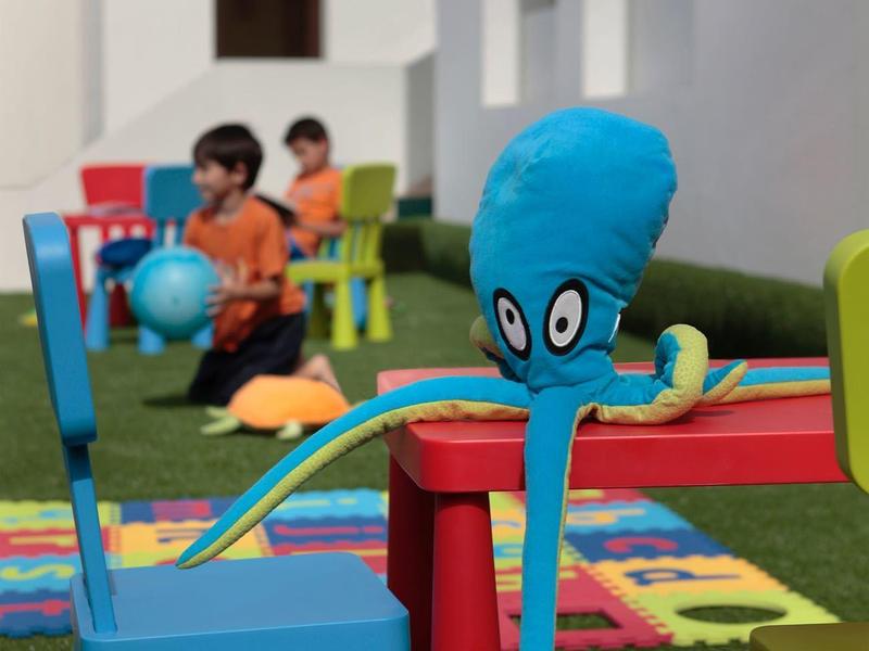 Bunter Spielbereich mit bunten Stühlen, Teppichen und einem blauen Tintenfisch-Charakter.