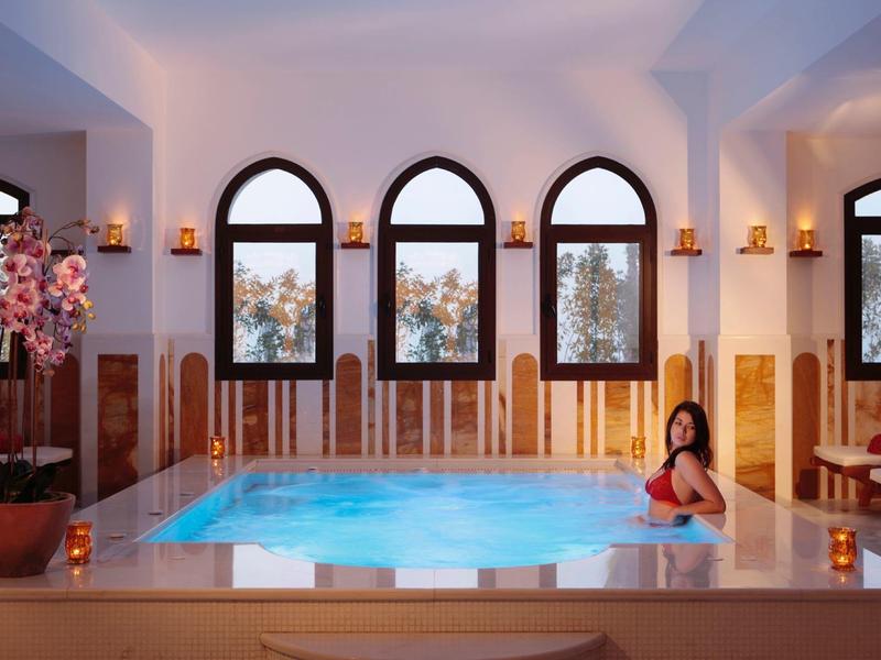 Innenraum mit großem Whirlpool, vier hohen Fenstern und entspannter Frau im roten Badeanzug.