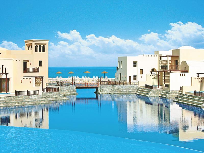 Weißes Gebäude mit Palmen am Meer, gepflasterter Weg, Sonnenuntergang im Hintergrund.