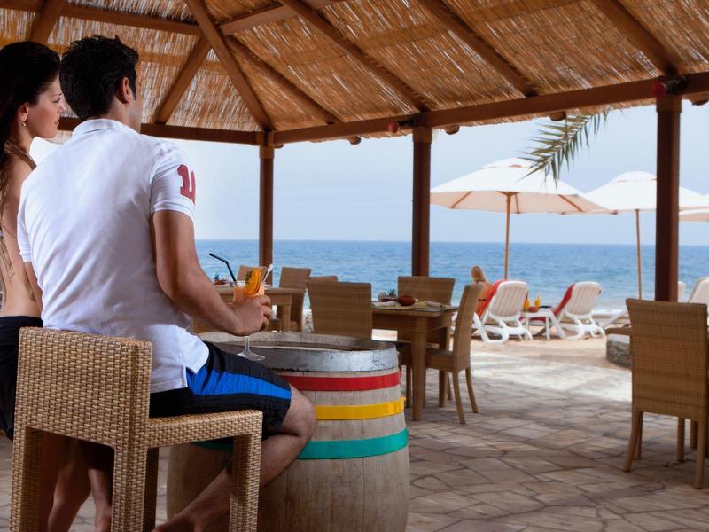 Paar sitzt unter Strohdach, blickt aufs Meer, bunte Fass-Tisch, Sandboden, liegestühle mit Sonnenschirm.