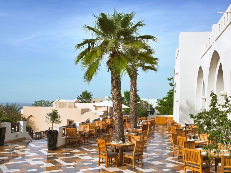Terrasse mit Holztischen, Stühlen, Palmen und weißem Gebäude unter blauem Himmel.