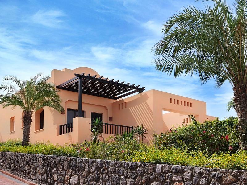 Flaches Haus in Erdtönen mit Pergola, umgeben von Palmen und grüner Bepflanzung bei blauem Himmel.