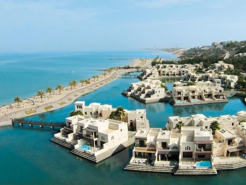 Inselanlage mit weißen Häusern auf Stegen, türkises Wasser und grüne Hügel im Hintergrund.