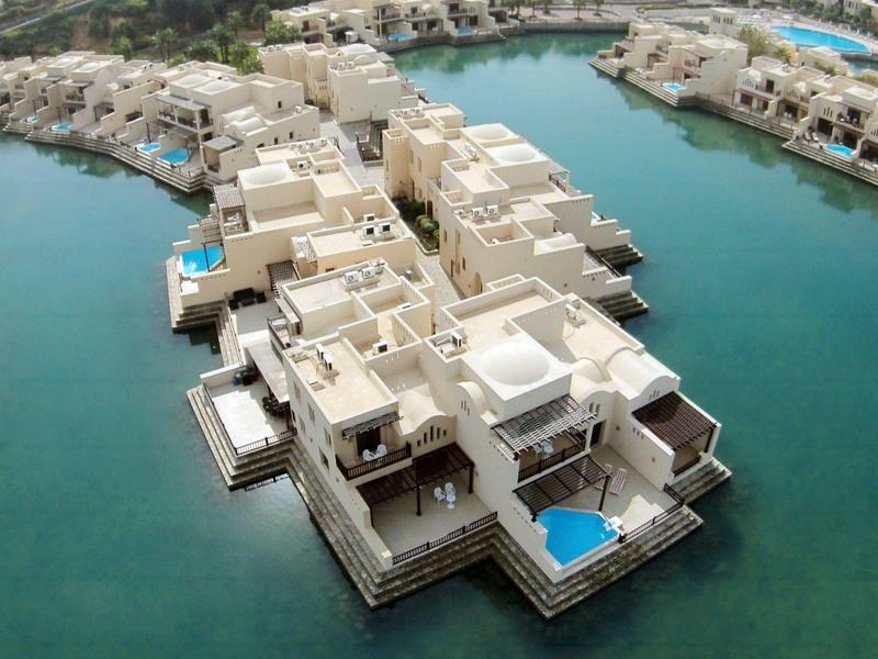 Luftaufnahme einer Wohnanlage mit weißen Gebäuden und blauen Pools, umgeben von türkisfarbenem Wasser.