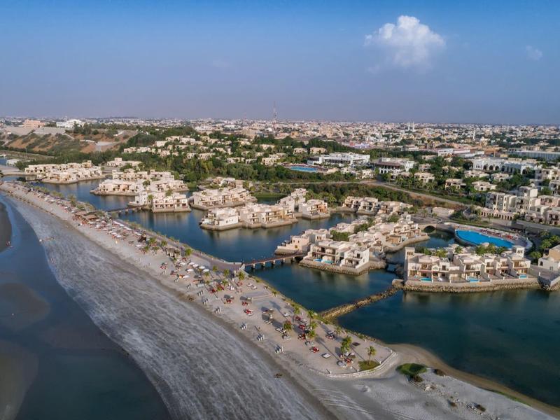 Küstenstadt mit Sandstrand, Wasserkanälen und dicht aneinander gebauten weißen Gebäuden bei klarem Himmel.