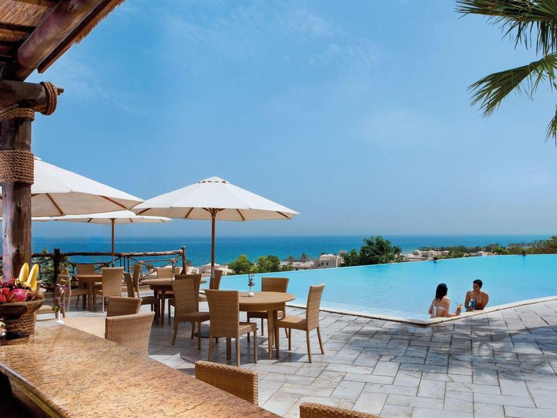 Infinity-Pool mit blauem Wasser, Terrassenmöbeln, Sonnenschirmen und Blick aufs Meer und Palmen.