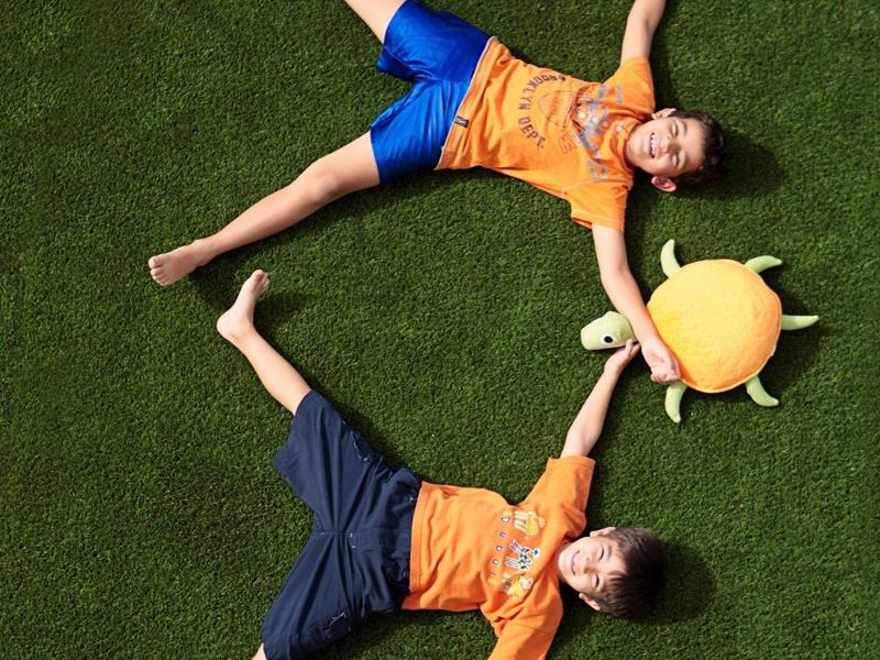 Zwei Kinder in orangenen Shirts liegen auf grünem Gras, greifen sich an die Hände, ein Sonnenspielzeug liegt daneben.