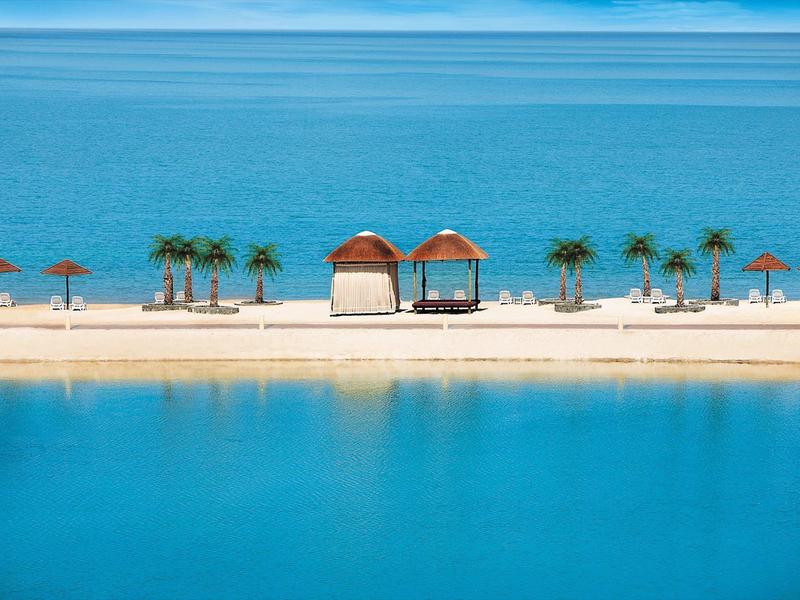 Strand mit Palmen, Sonnenschirmen und zwei kleinen Holzhütten vor ruhigem, blauem Meer.