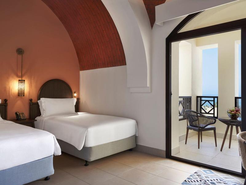 Helles Doppelzimmer mit zwei Betten, Holzbalken, und Balkon mit Tisch und zwei Stühlen.