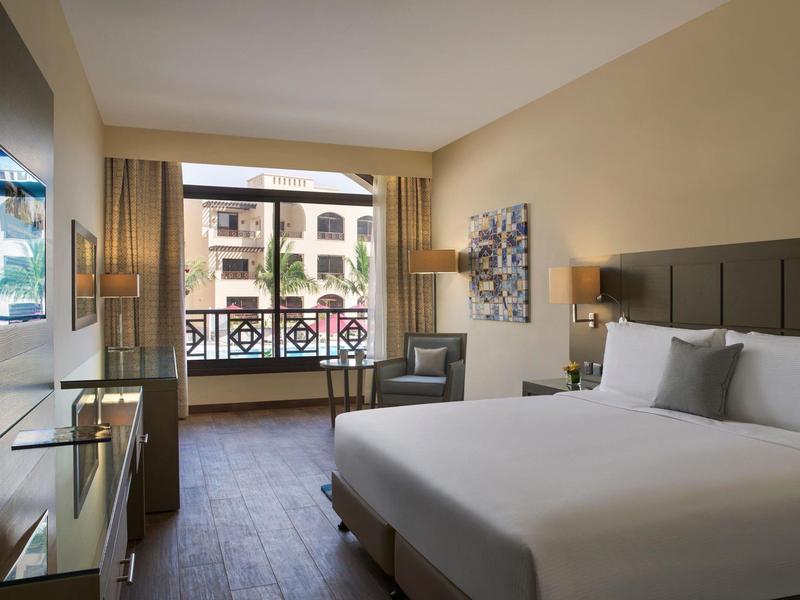 Helles Hotelzimmer mit großem Bett, Sessel, Schreibtisch und Balkon mit Blick auf Gebäude.