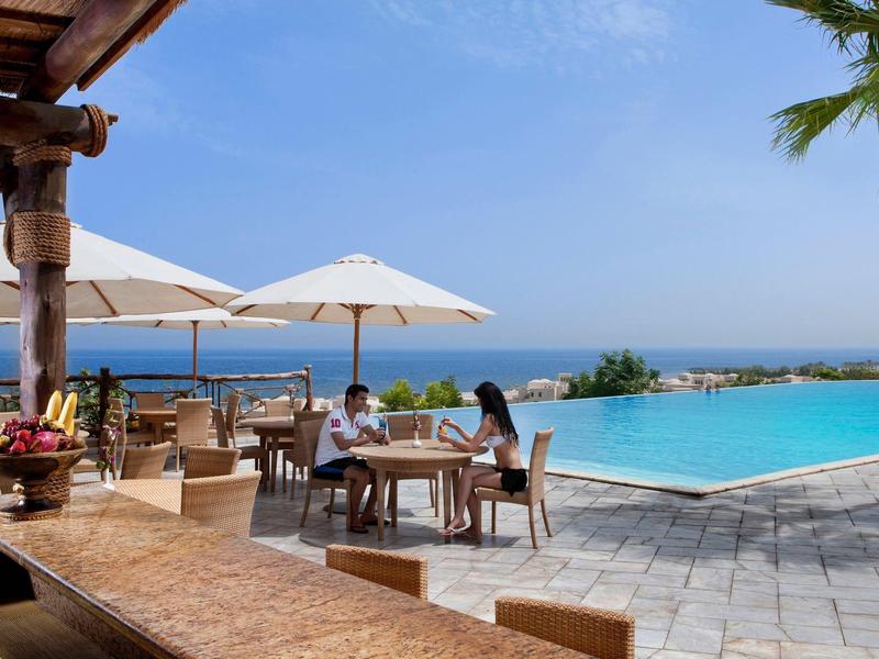 Terrasse mit Pool, weißen Sonnenschirmen und Meerblick, zwei Personen sitzen am Tisch.