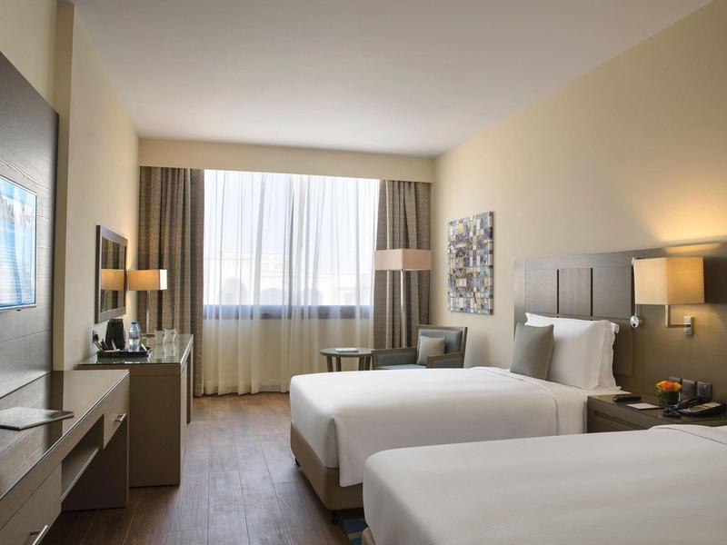 Hotelzimmer mit zwei weißen Betten, großen Fenster, Vorhängen, Lampen und moderner Einrichtung.