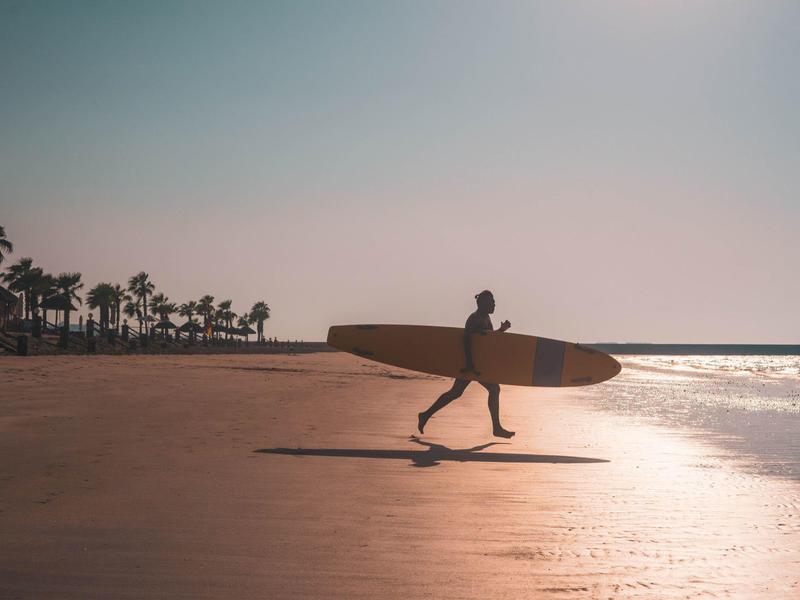 Ein Surfer läuft mit einem Surfbrett am Sonnenuntergangsstrand entlang, Palmen sind im Hintergrund.