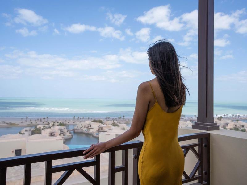 Eine Frau in gelbem Kleid blickt von einem Balkon auf einen sonnigen Strand und das Meer.