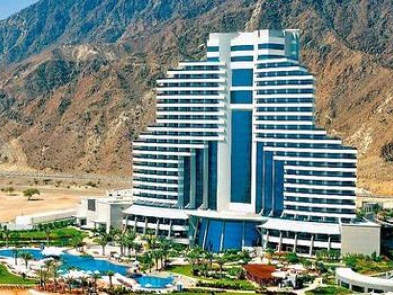 Großes mehrstöckiges Hotel mit Terrassen vor felsigen Bergen und trockenem, braunem Gelände.