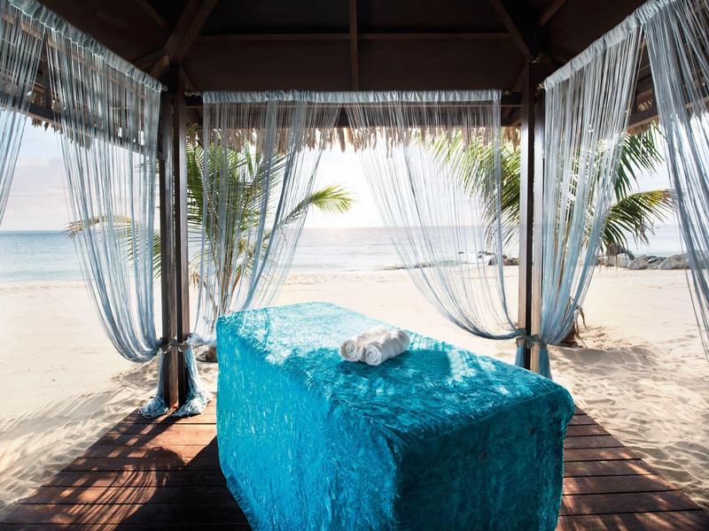 Massagebereich mit türkisfarbener Liege unter Pavillon am Strand mit Blick aufs Meer