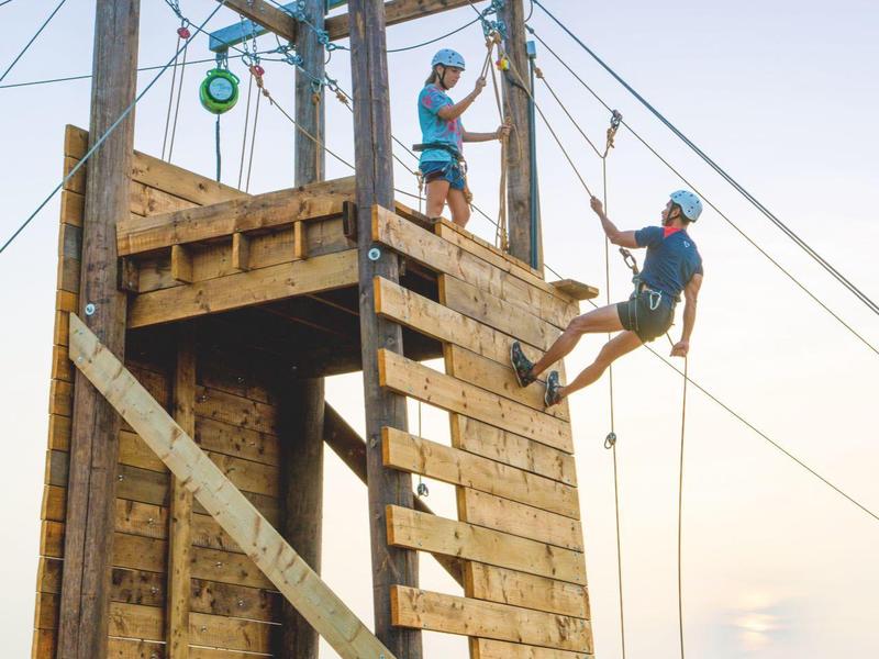 Personen erklimmen einen Kletterturm mit Seil im Freien bei klarem Himmel.