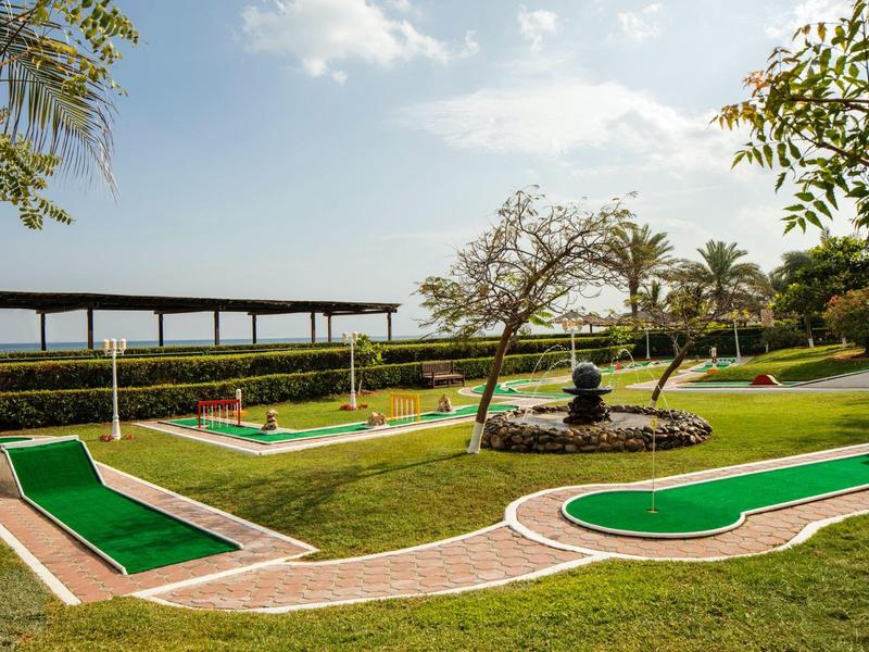 Minigolfanlage mit grünen Bahnen, Springbrunnen, Bäumen und überdachtem Bereich im Hintergrund