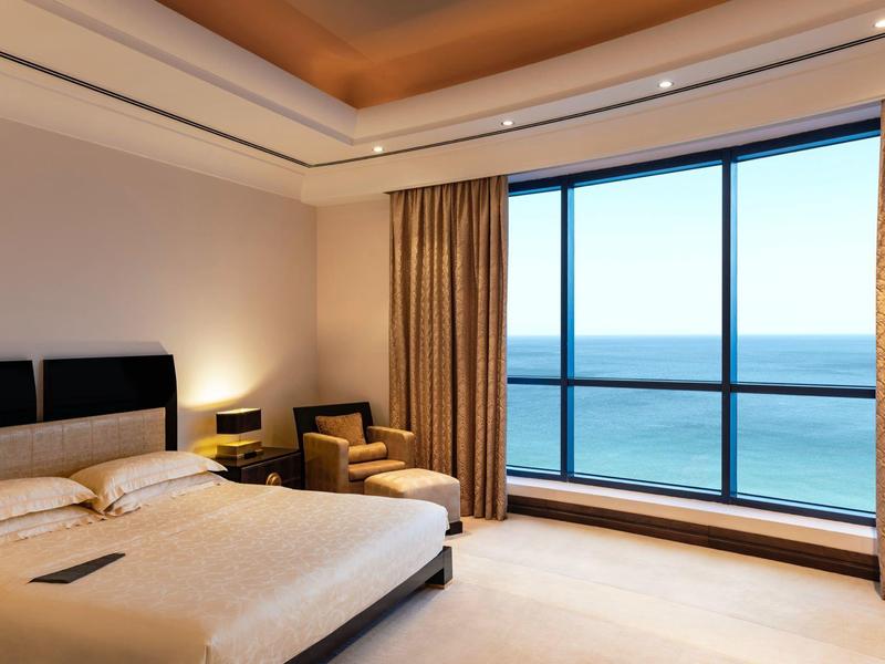 Modernes Hotelzimmer mit großem Bett, Sesseln und Panoramablick aufs Meer durch raumhohe Fenster.