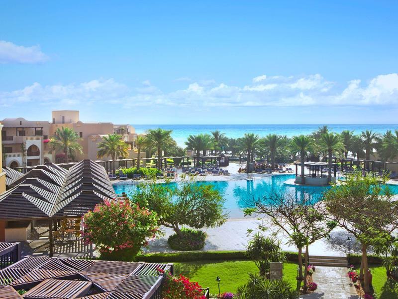 Blick auf einen Hotelpool mit Palmen und Meer im Hintergrund bei sonnigem Wetter
