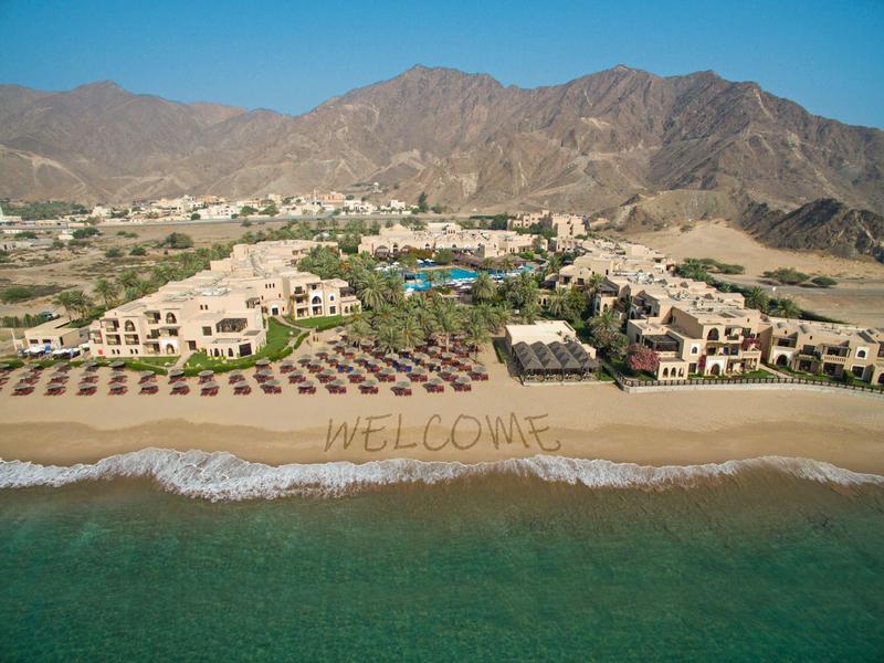 Strand mit 'WELCOME' im Sand, Sonnenschirmen und Hotelgebäuden vor Gebirgslandschaft