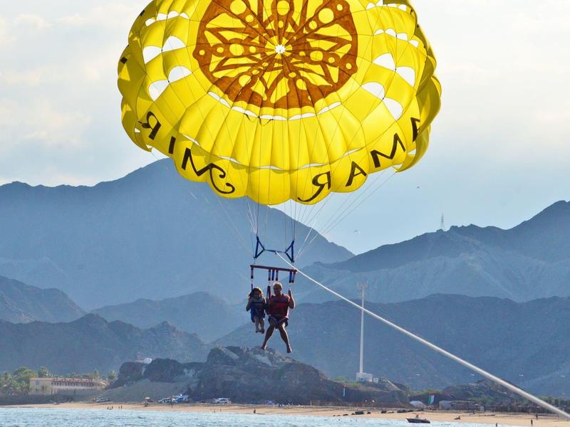 Zwei Personen beim Parasailing mit gelbem Fallschirm über dem Wasser vor Bergen.
