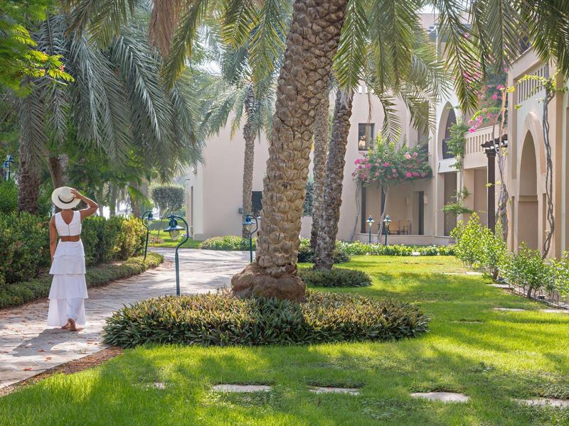 Eine Frau in einem weißen Kleid und Hut geht durch eine Gartenanlage mit Palmen neben einem Hotelgebäude.