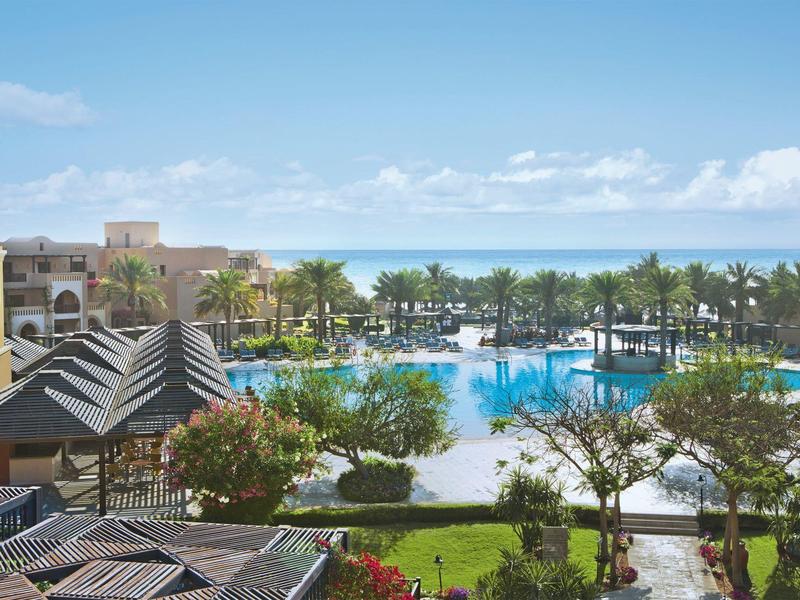 Blick auf einen Hotelpool mit Palmen, Grünflächen und Meer im Hintergrund bei klarem Himmel.
