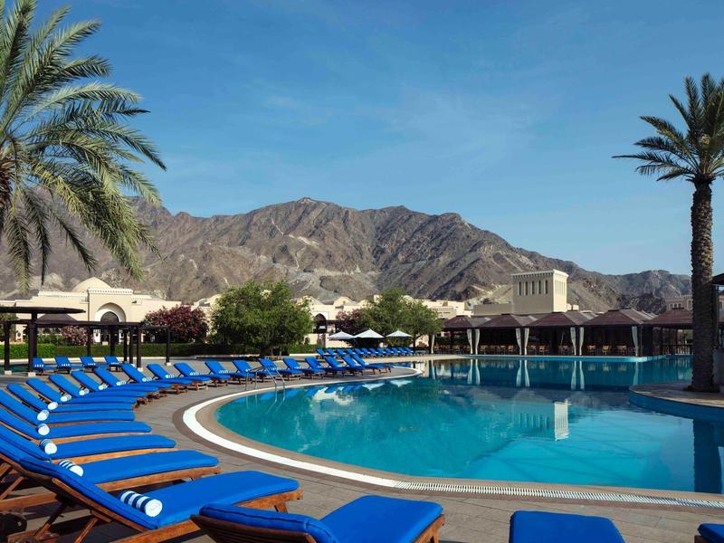 Runder Hotelpool mit blauen Liegestühlen, Palmen und Bergkulisse im Hintergrund.