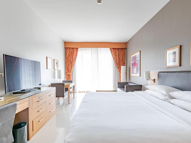 Modernes Hotelzimmer mit Doppelbett, kleinem Schreibtisch, Fernseher und großen Fenstern mit Vorhängen.