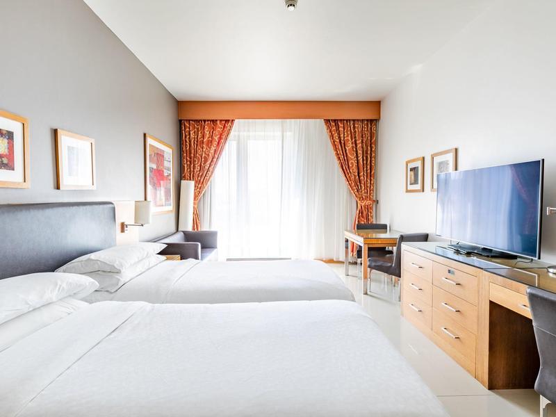 Helles Hotelzimmer mit zwei Betten, Fernseher, Schreibtisch, Stuhl und großen Fenstern mit orangen Vorhängen.
