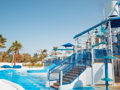 Wasserpark mit blauen Rutschen, Treppen und Palmen unter klarem Himmel.