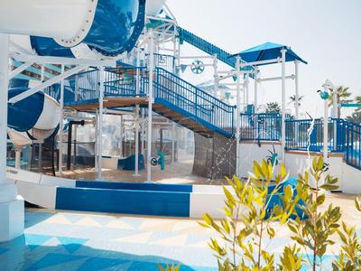 Buntes Wasserspielplatz mit blauen Rutschen, Treppen und Palmen im Sonnenlicht.