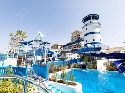 Wasserpark mit blauen Rutschen, Becken und weiß-blau gestreiftem Turm unter klarem Himmel.