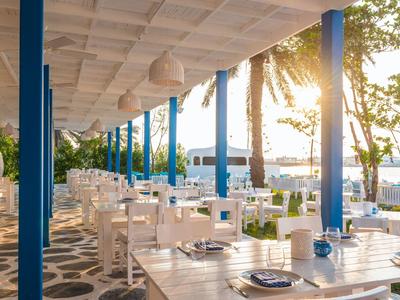 Offenes Restaurant mit weißen Tischen, blauen Säulen und Blick auf Palmen und Meer bei Sonnenuntergang.