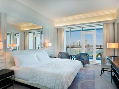 Modernes Schlafzimmer mit weißem Bett, großen Fenstern, zwei Sesseln und Schreibtisch.