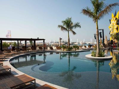 Rundlicher Pool mit Liegestühlen, Palmen und klar blauem Himmel in einer Hotelanlage.