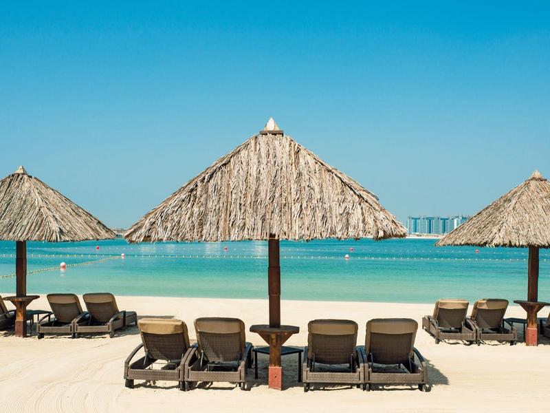 Drei Strohdach-Schirme mit Liegestühlen auf weißem Sandstrand und türkisfarbenem Meer darunter.