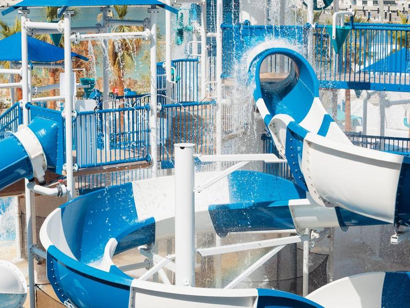 Wasserpark mit blauen und weißen Wasserrutschen, Wassereffekten und Sonnenschirmen im Hintergrund.