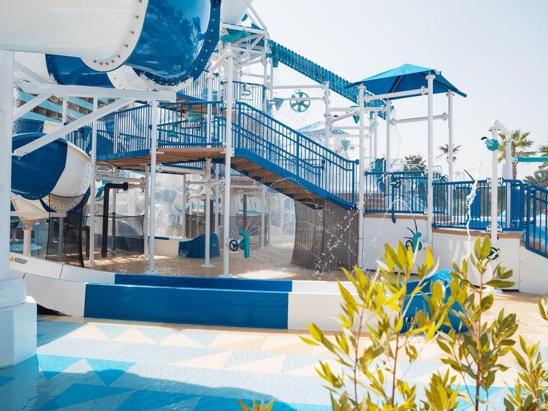 Buntes Wasserspielplatz mit blauen Rutschen, Treppen und Palmen im Sonnenlicht.