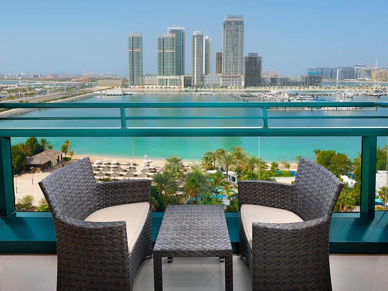 Balkon mit grauen Korbsesseln und Tisch, Blick auf blaue Bucht und hohe Gebäude am Horizont.