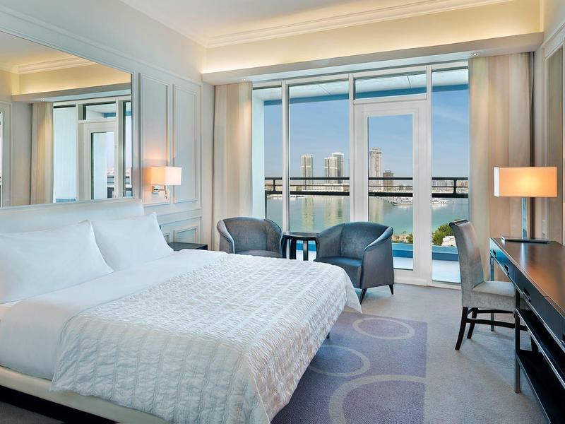 Modernes Hotelzimmer mit großem Bett, zwei Sesseln, Schreibtisch und Blick auf einen Balkon mit Wasserblick.