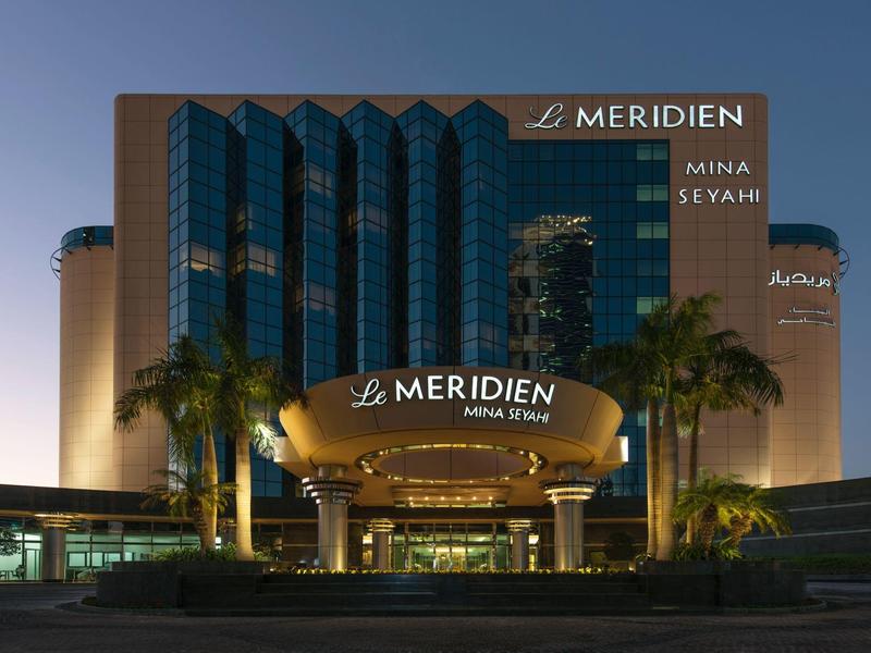 Moderner Hotelkomplex mit gläserner Fassade, Logo "Le Meridien" und Palmen davor bei Dämmerung.