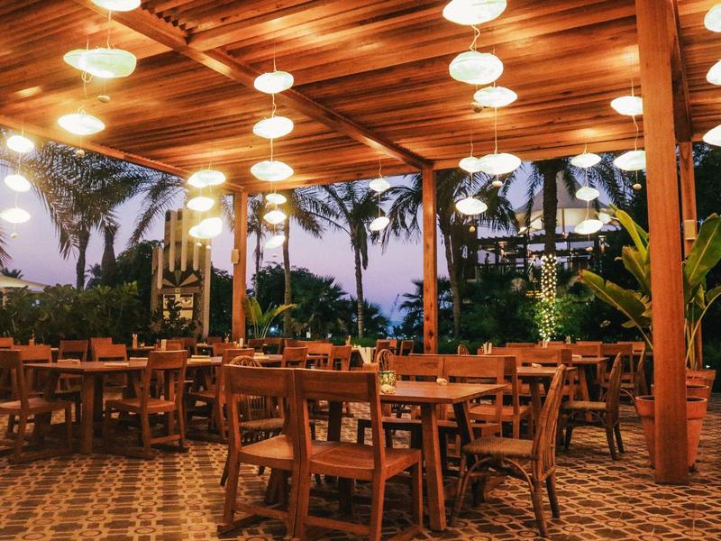 Freiluftrestaurant mit Holzdecke, hellen Lampen und vielen Tischen, umgeben von Palmen.