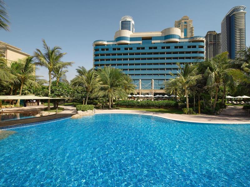 Großer blauer Pool vor moderner Hotelanlage mit Palmen und Liegestühlen an sonnigem Tag.