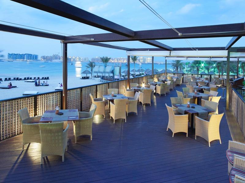 Offene Terrasse mit hellen Korbsesseln und Tischen, Blick auf ruhiges Wasser und entfernte Gebäude.